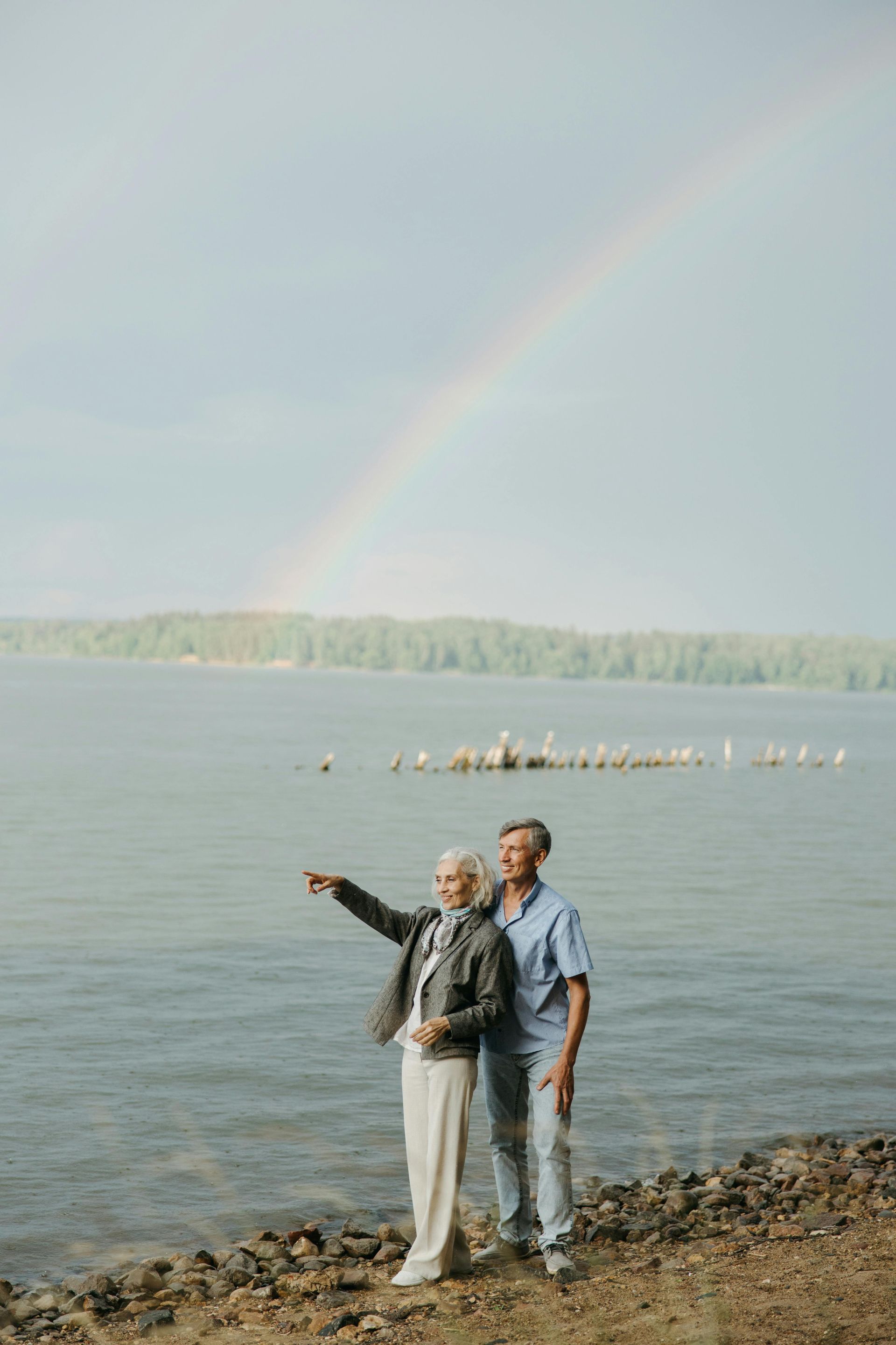 Two people standing by a lake, one pointing across the water under a faint rainbow