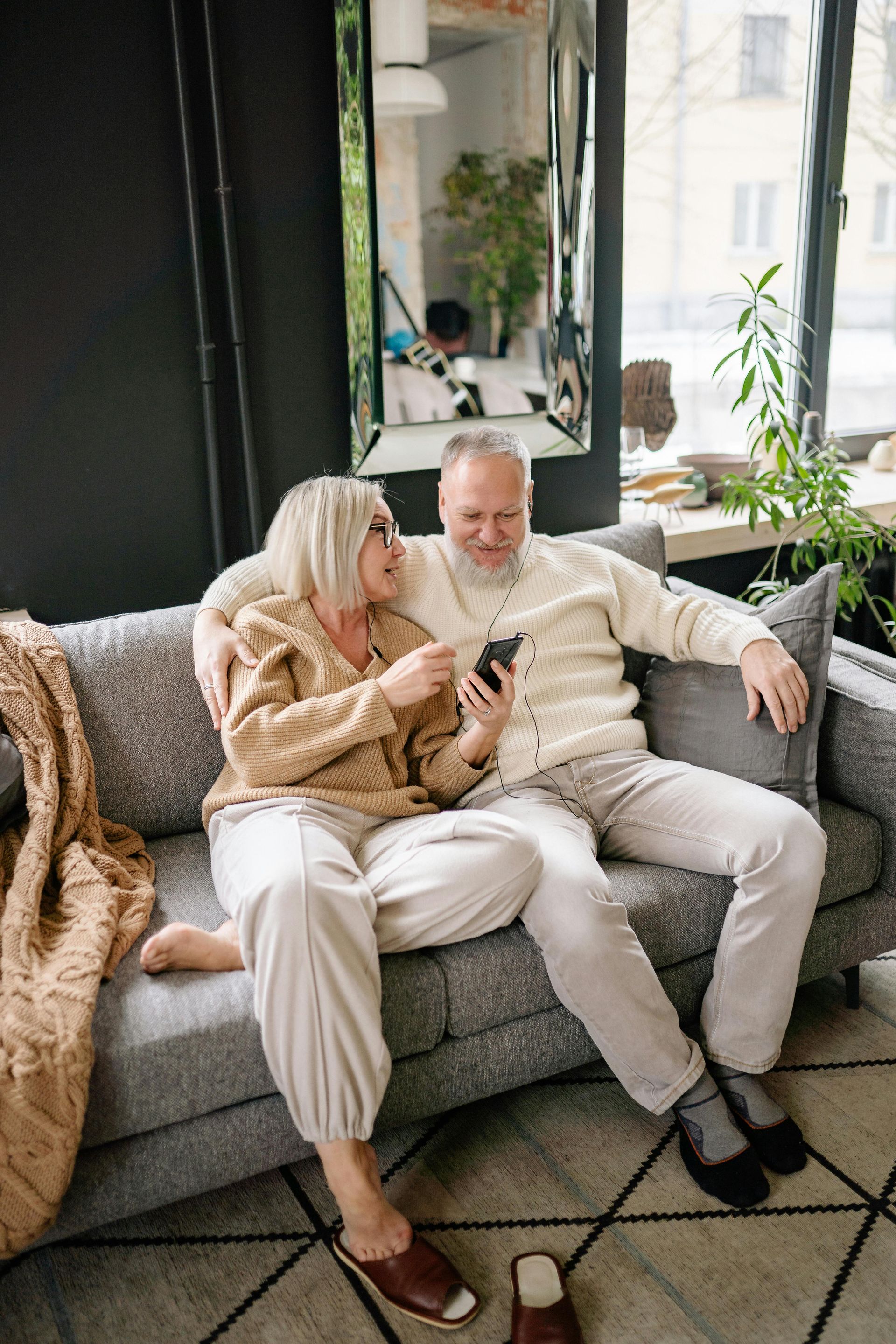 Couple cuddling on a gray sofa in a bright living room with plants and a large mirror.