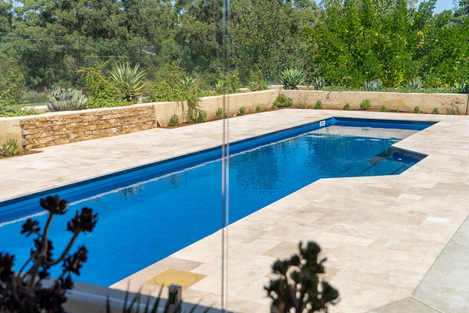 Rectangular blue swimming pool surrounded by beige tiles and stone wall, with greenery in background.