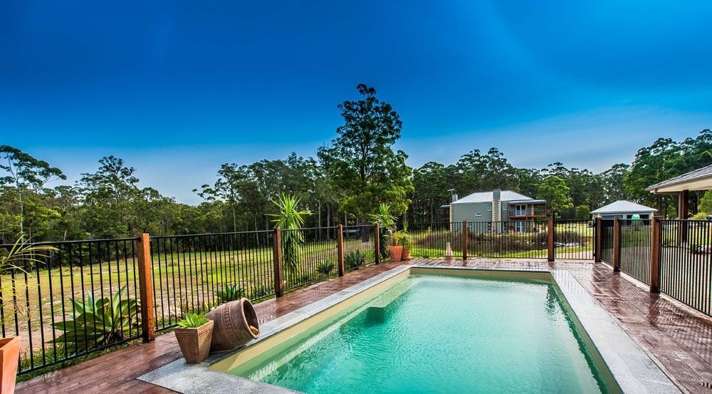 A Large Swimming Pool is Surrounded by a Fence and Trees — Gibson Family Pools Pty Ltd In Port Macquarie, NSW