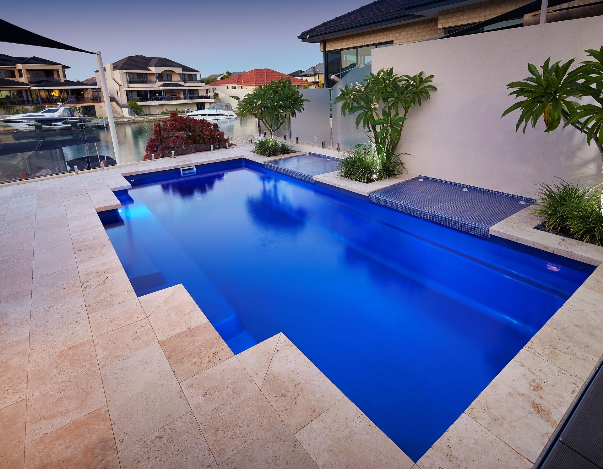 Blue rectangular pool in backyard setting with stone patio and adjacent buildings.