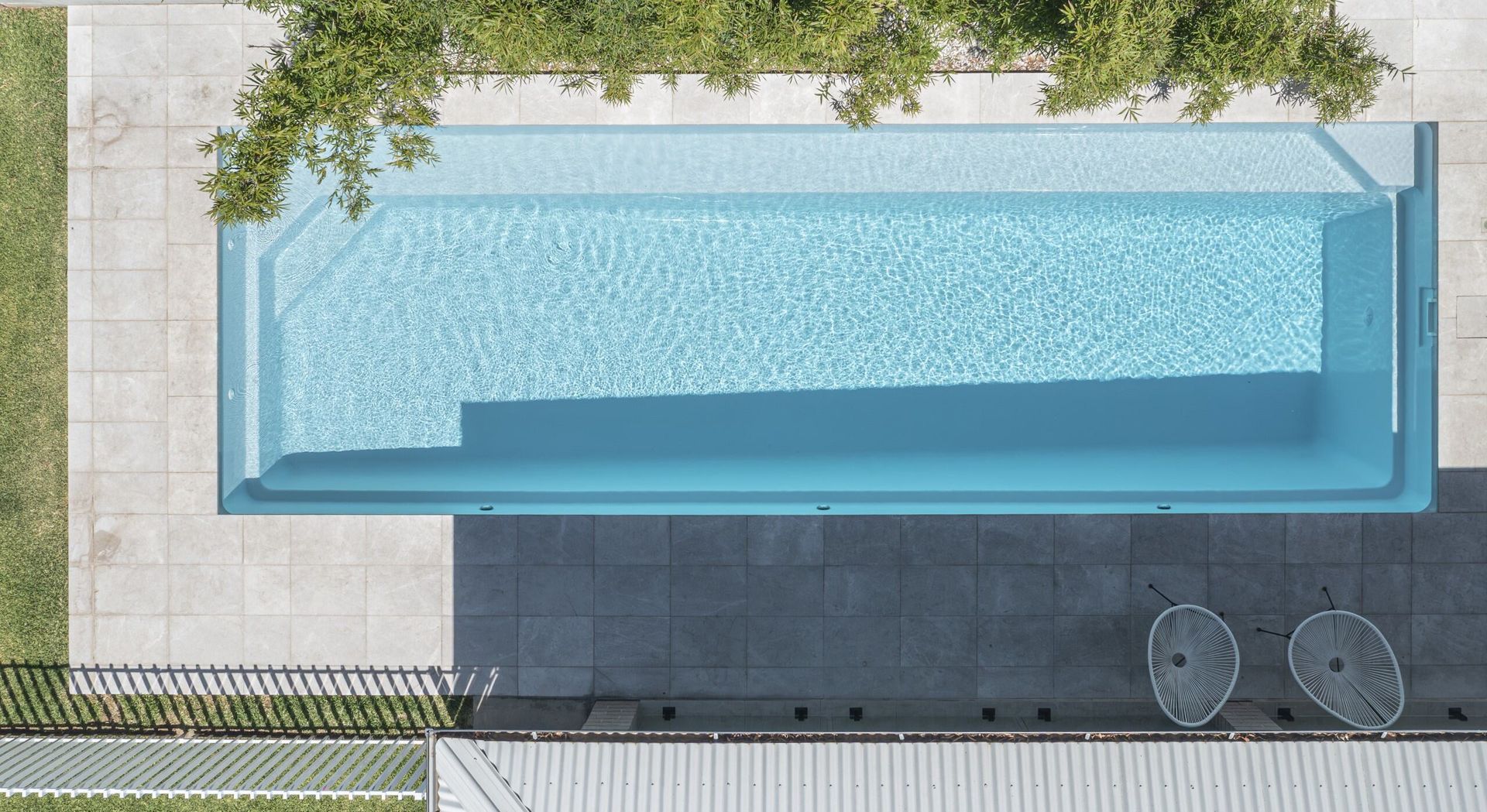 Overhead view of a rectangular swimming pool with steps, surrounded by patio, grass, and a fence.