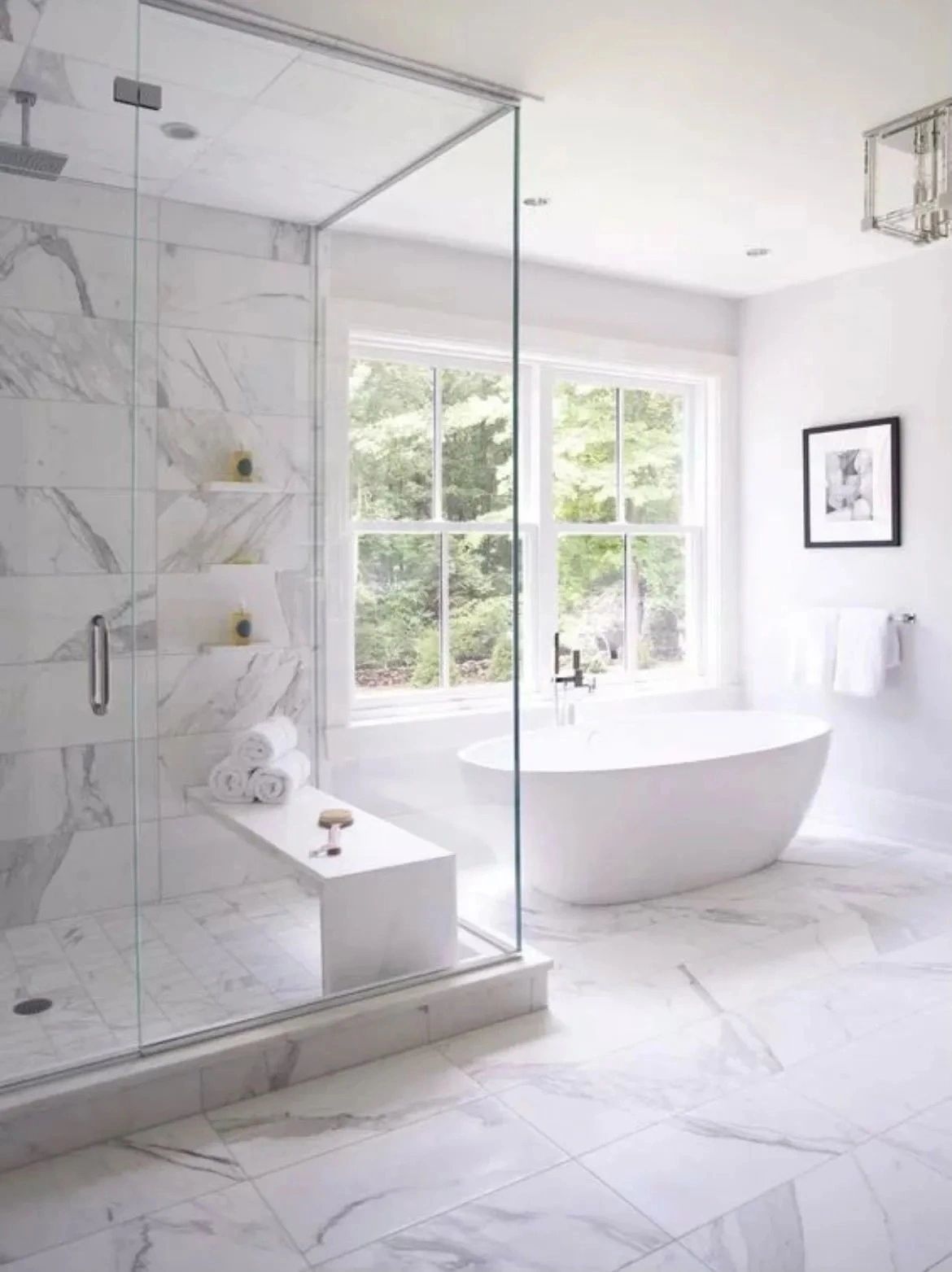 Bright marble bathroom with glass shower, freestanding tub, and large window overlooking greenery