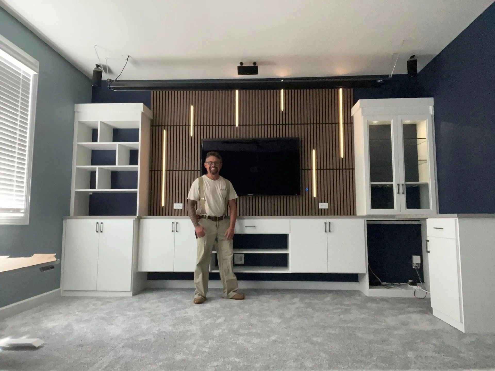 Person standing in a modern living room with a TV wall, white cabinets, and gray carpet
