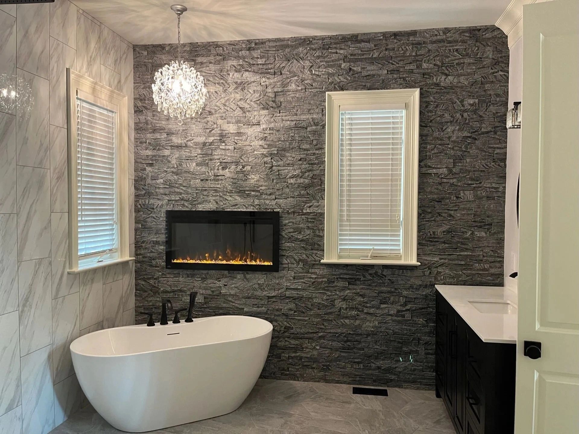 Modern bathroom with freestanding tub, wall fireplace, dark textured tiles, and two windows