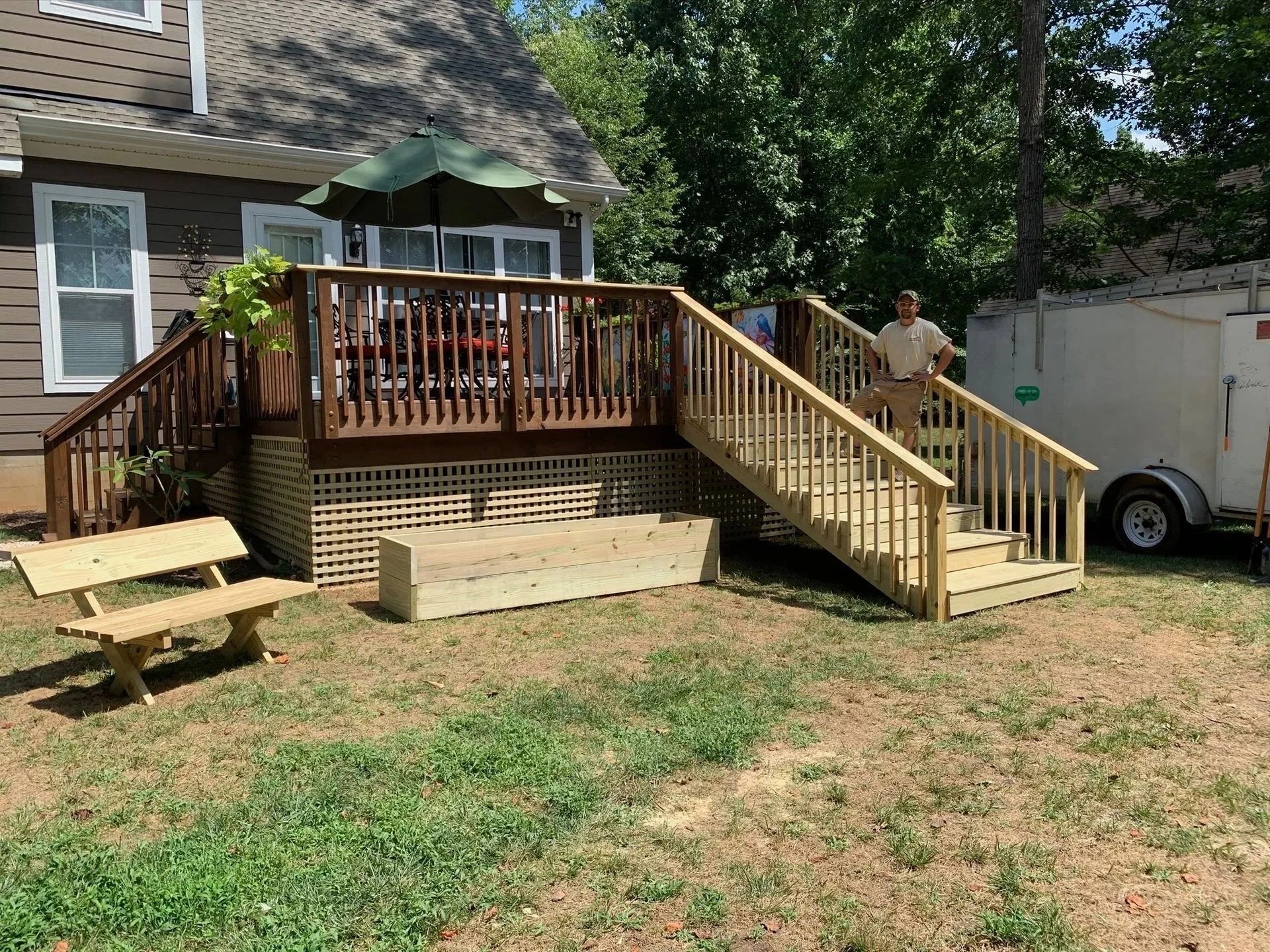 Backyard deck with stairs, wooden railing, table, and white trailer beside a house