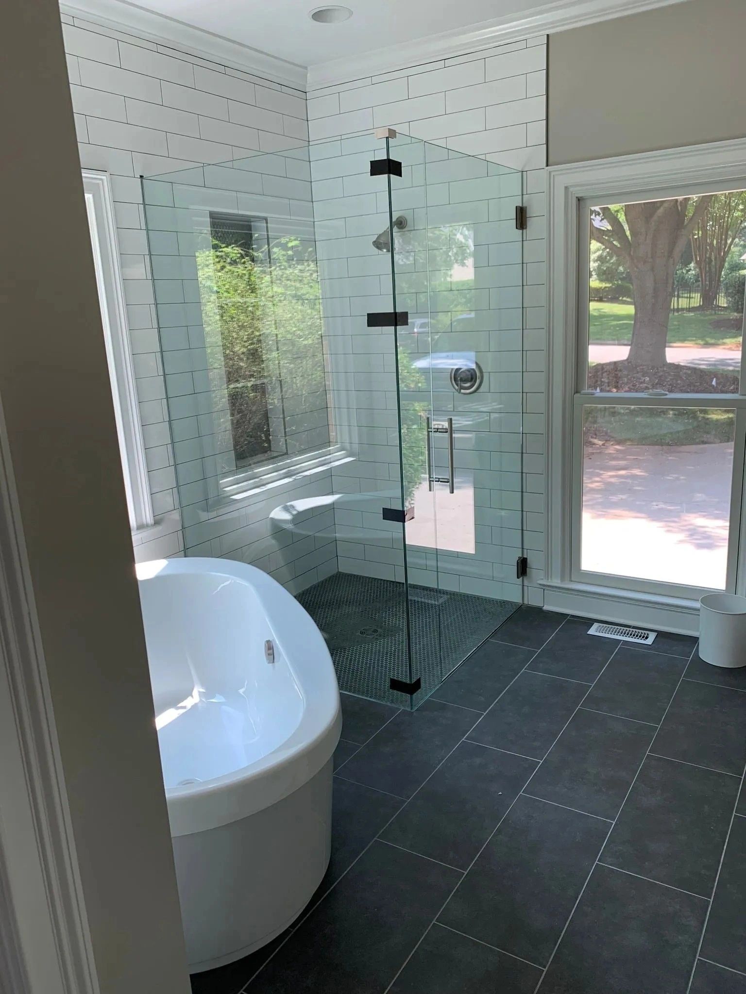 Modern bathroom with a freestanding tub, glass shower, black tile floor, and large windows overlooking trees