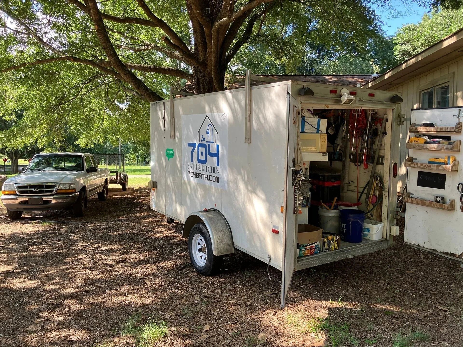 White utility trailer with open side door under a large tree, parked beside a house and truck in a yard.