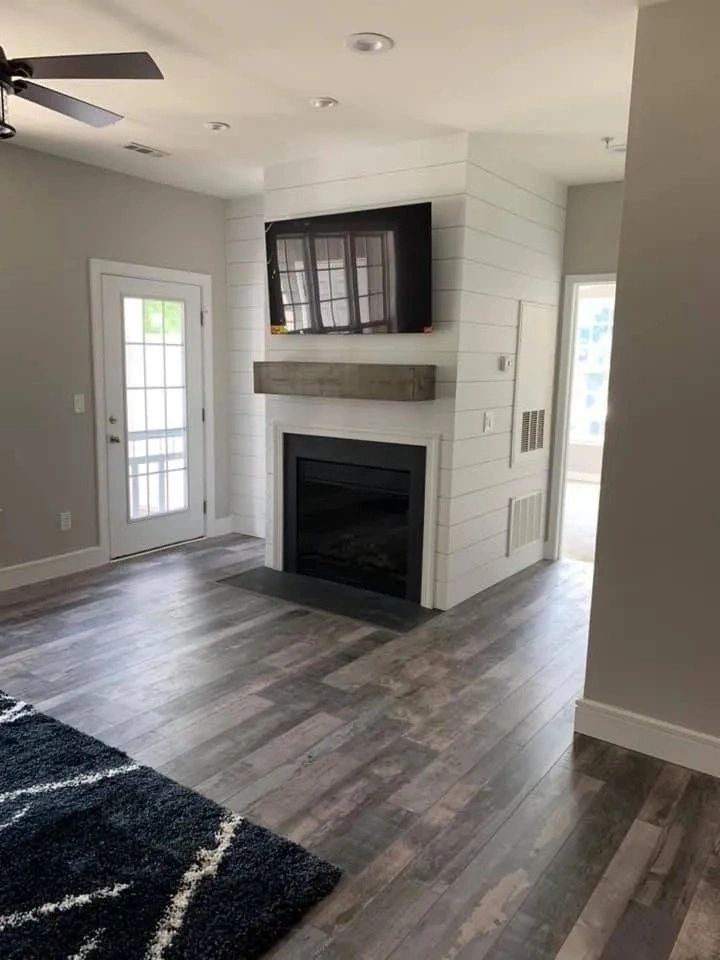 Living room with white shiplap fireplace, mounted TV, gray wood floors, and a glass door to the left