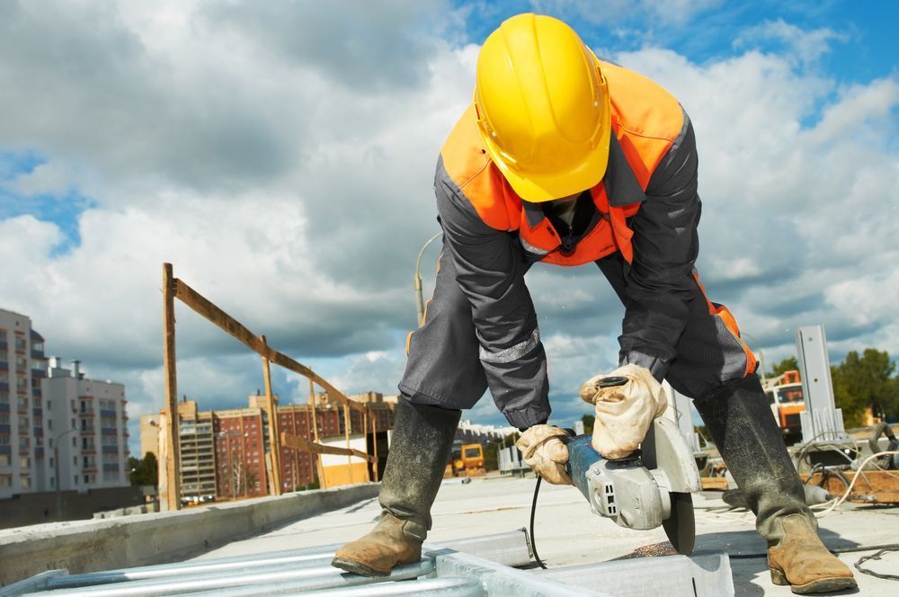Construction worker in orange vest and yellow hard hat using a power saw on a rooftop.
