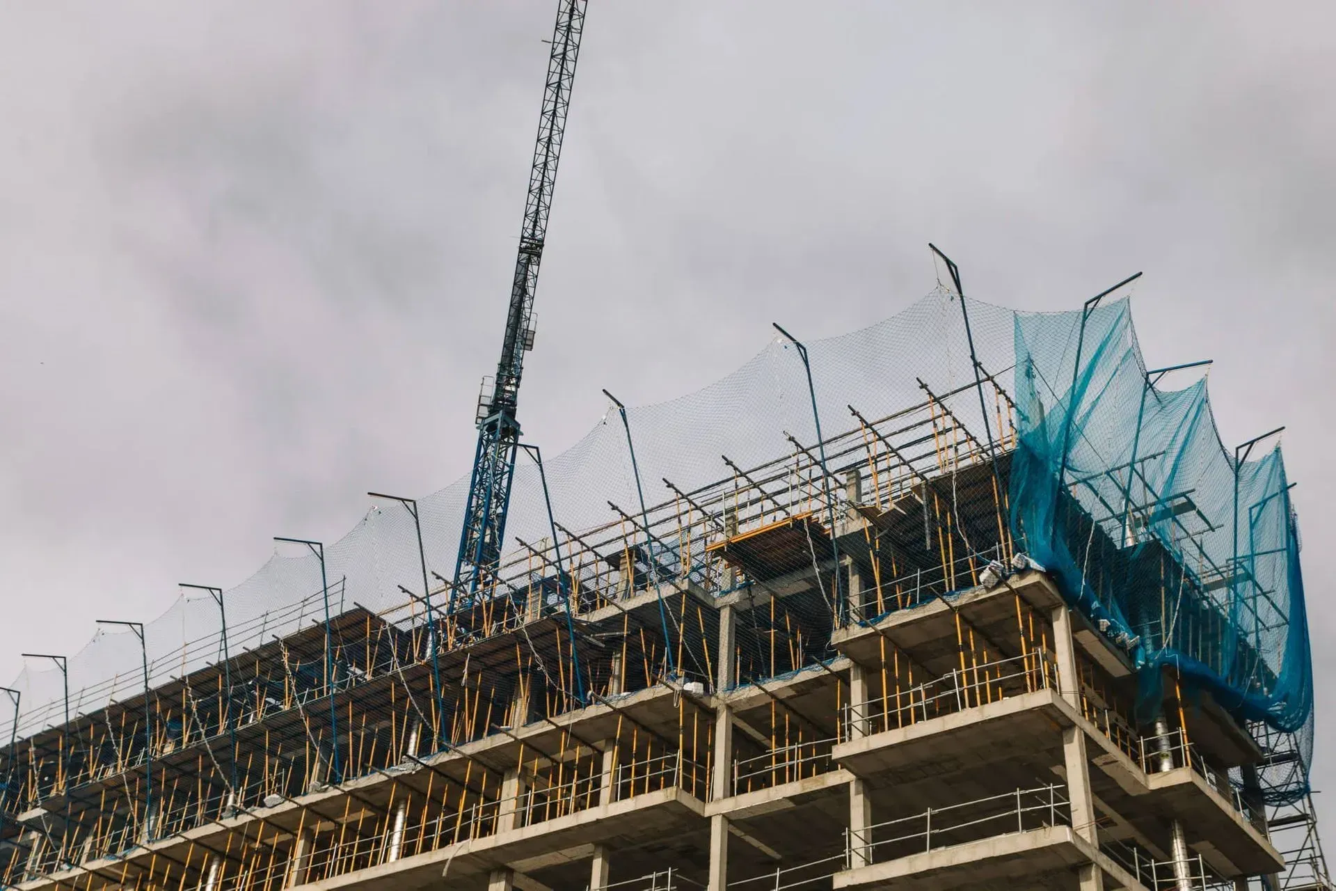 Partially built concrete structure with cranes and blue safety netting under a cloudy sky Partially built concrete structure with cranes and blue safety netting under a cloudy sky