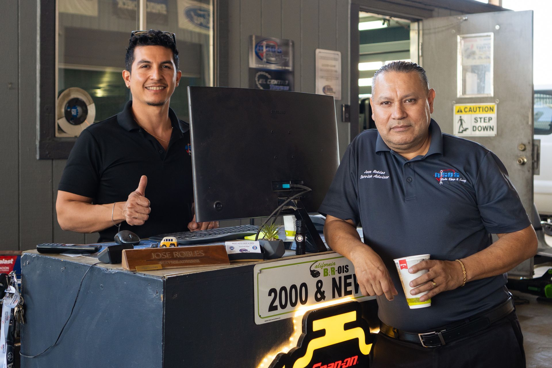 Two men at a repair shop counter. One gives a thumbs up. The other holds a cup.