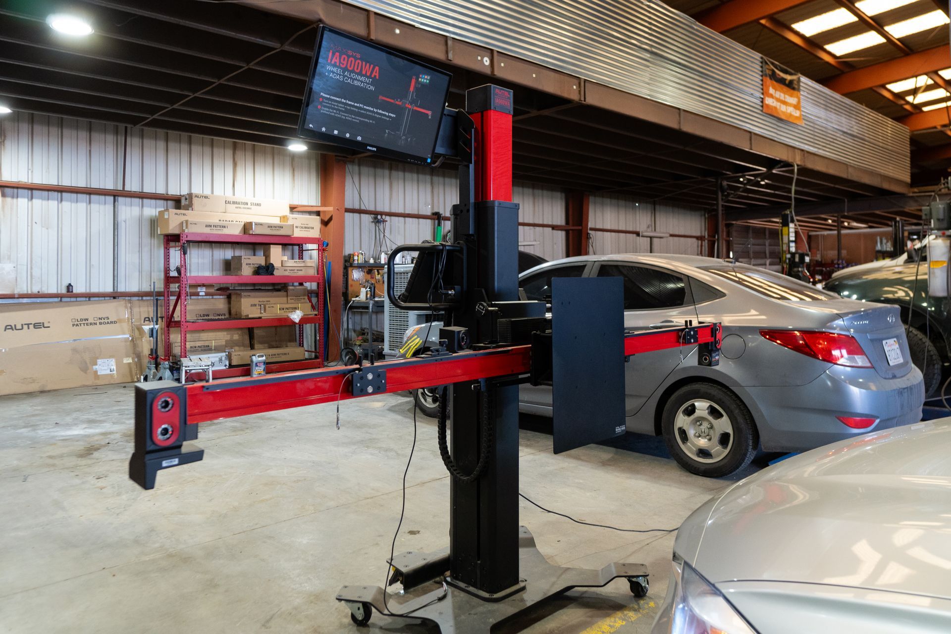 Wheel alignment machine in a garage, gray car, red and black equipment, shelving in the background.