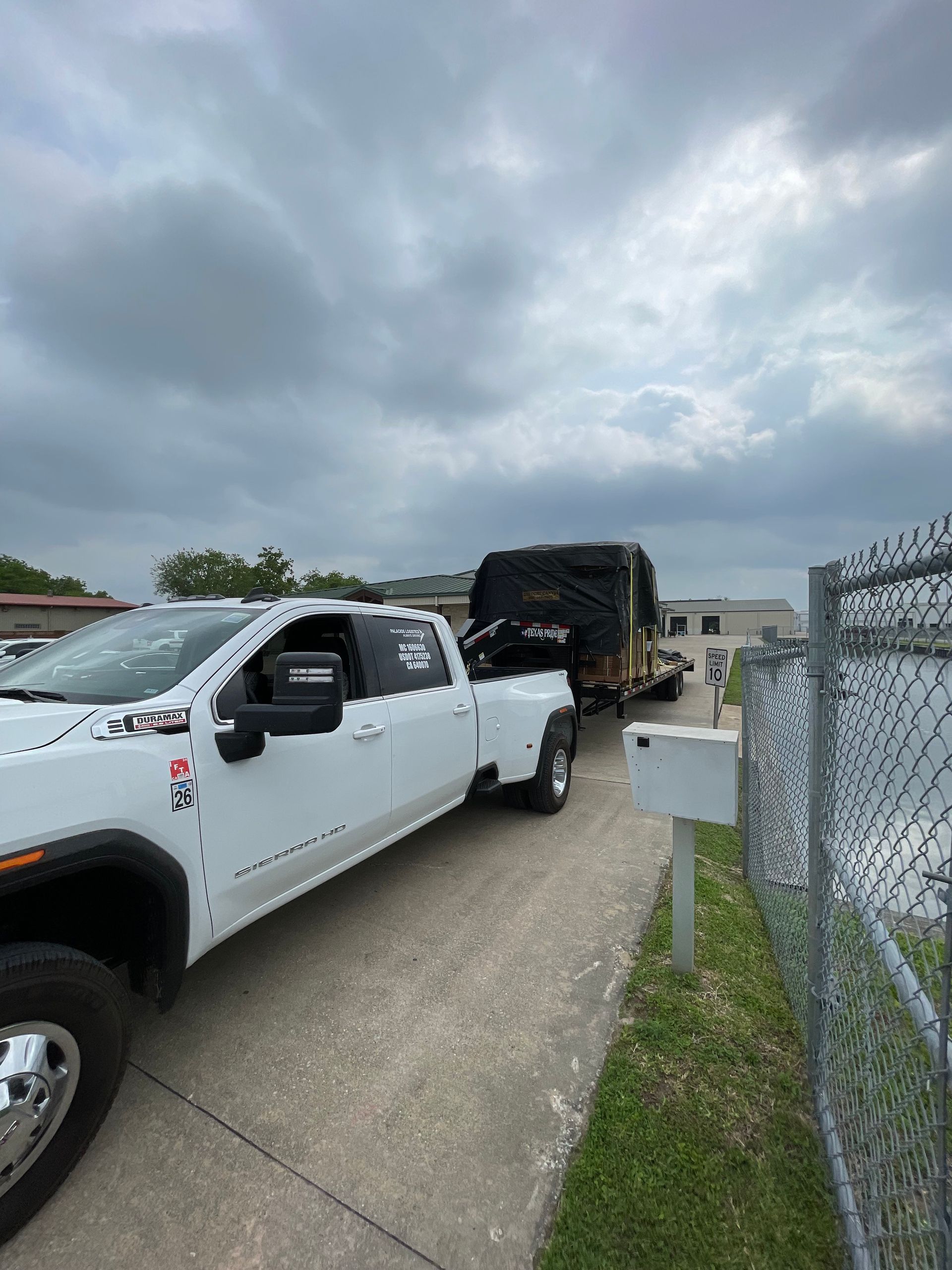 White truck towing a trailer with a large, covered object. Cloudy sky, next to a fence.