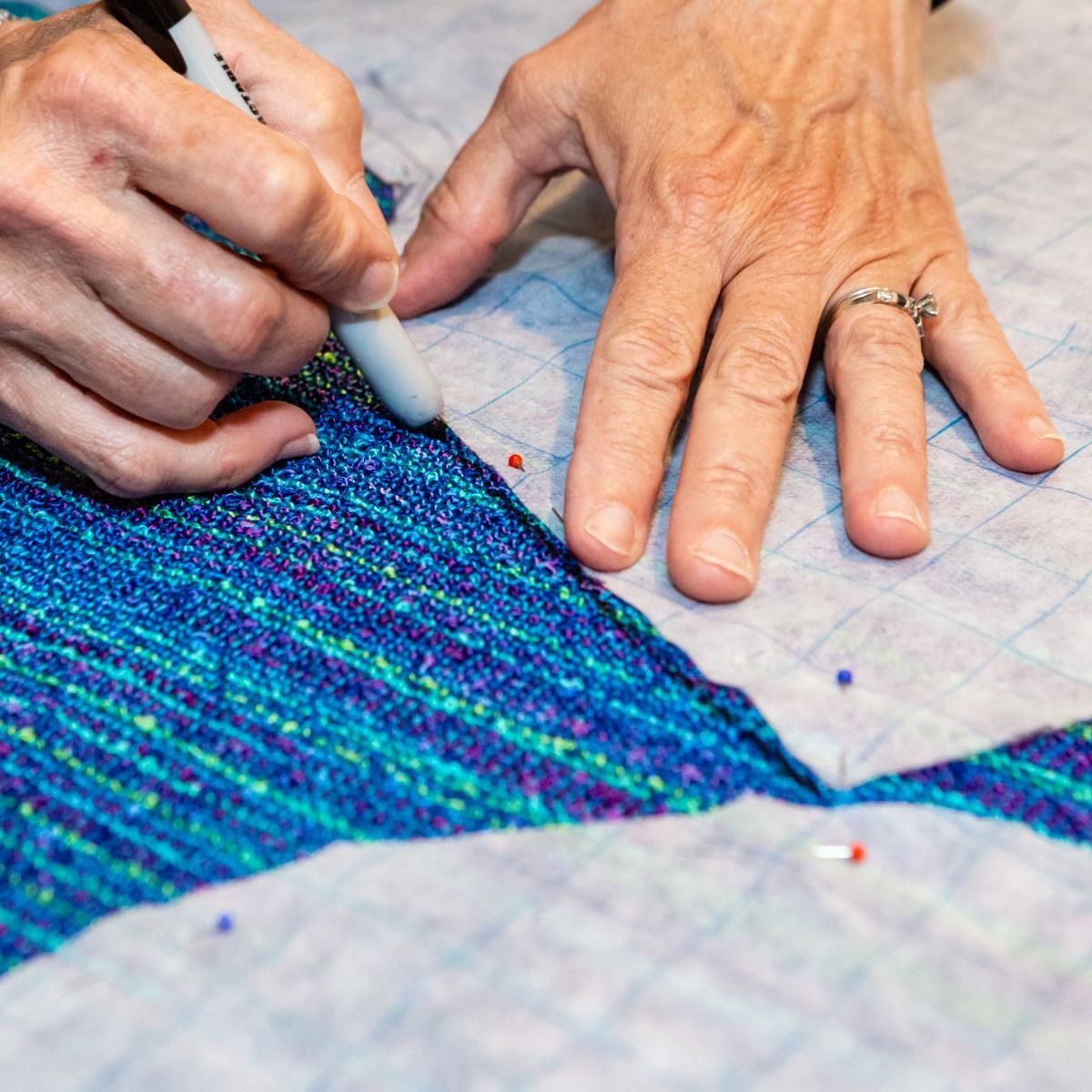 Hands pinning blue fabric while cutting a colorful woven textile pattern