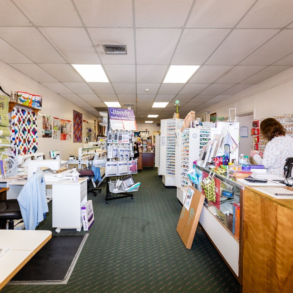 Small craft store aisle with white shelves, colorful supplies, and a person working at the counter