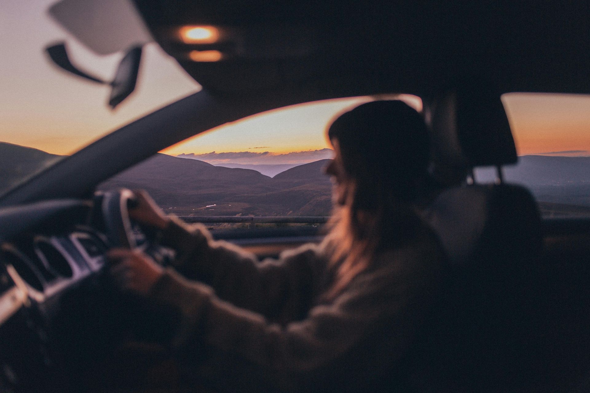 A woman is driving a car at sunset with mountains in the background.