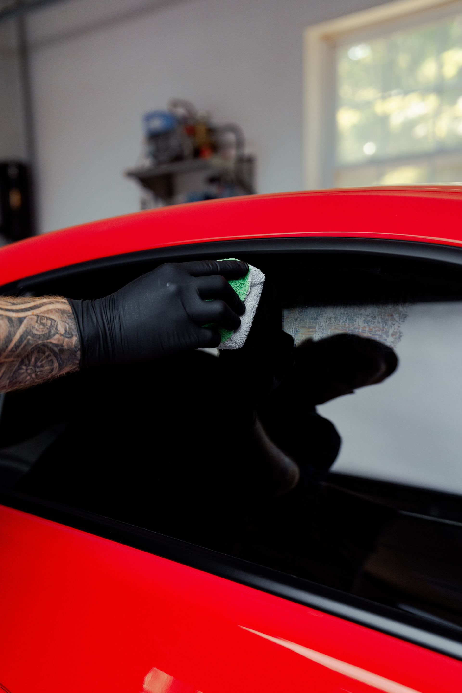 Person wearing black gloves cleaning a red car's window with a green sponge in a garage.