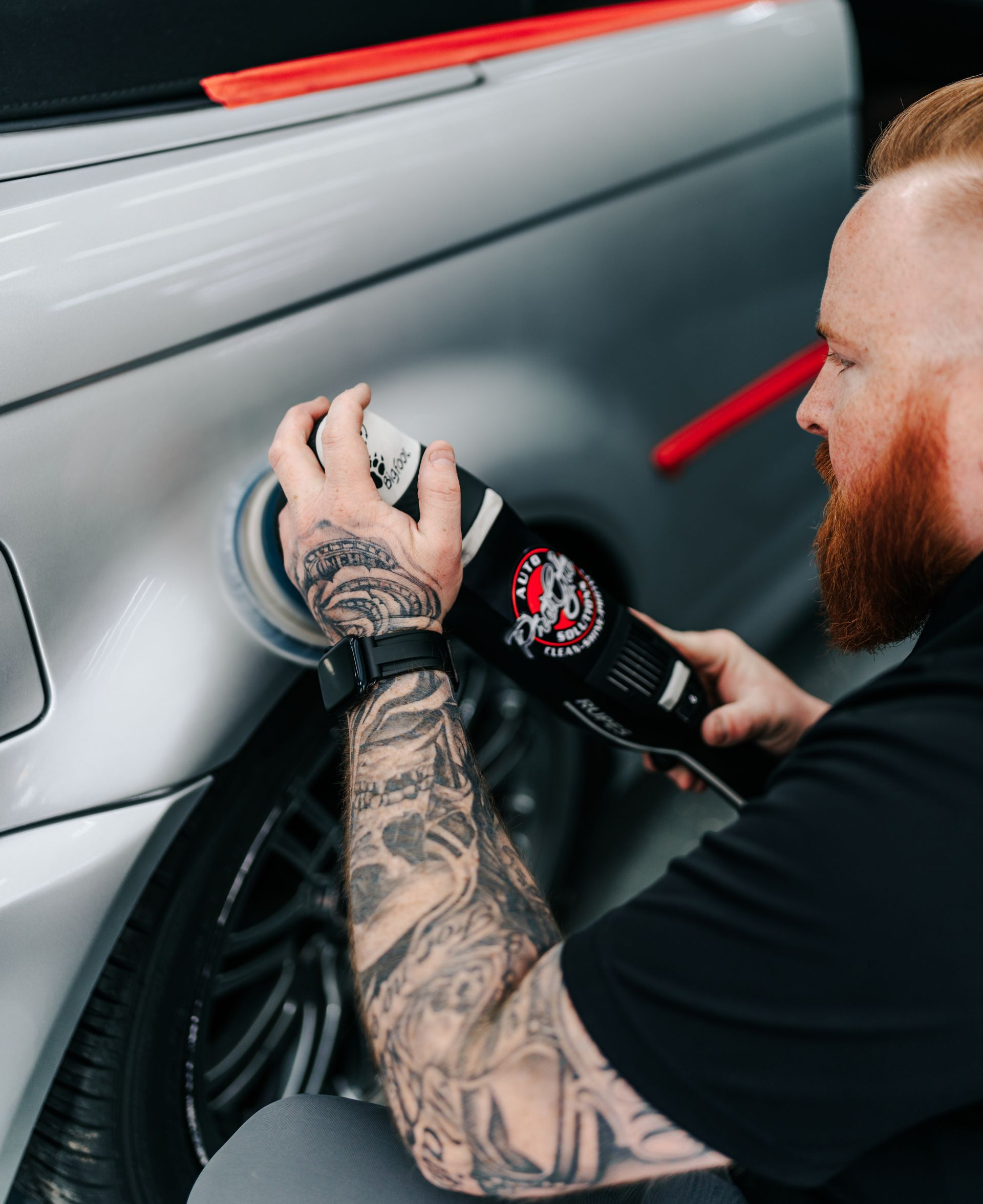 Person wearing black gloves polishes a car's black hood with a red and black machine in a garage.