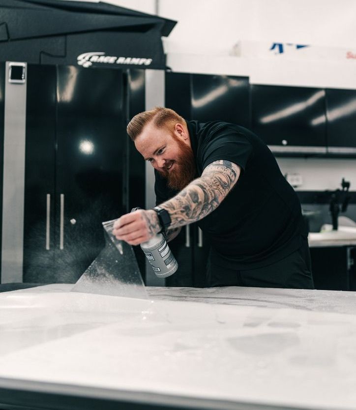 Man with a red beard sprays a surface, possibly applying a film in a shop.