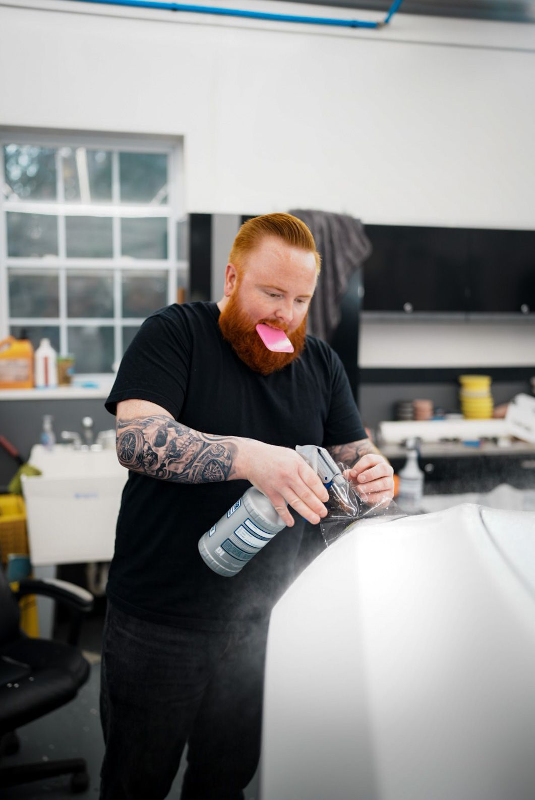 Man with red beard and tattoos spraying a car in a garage. Note stuck on his mouth.