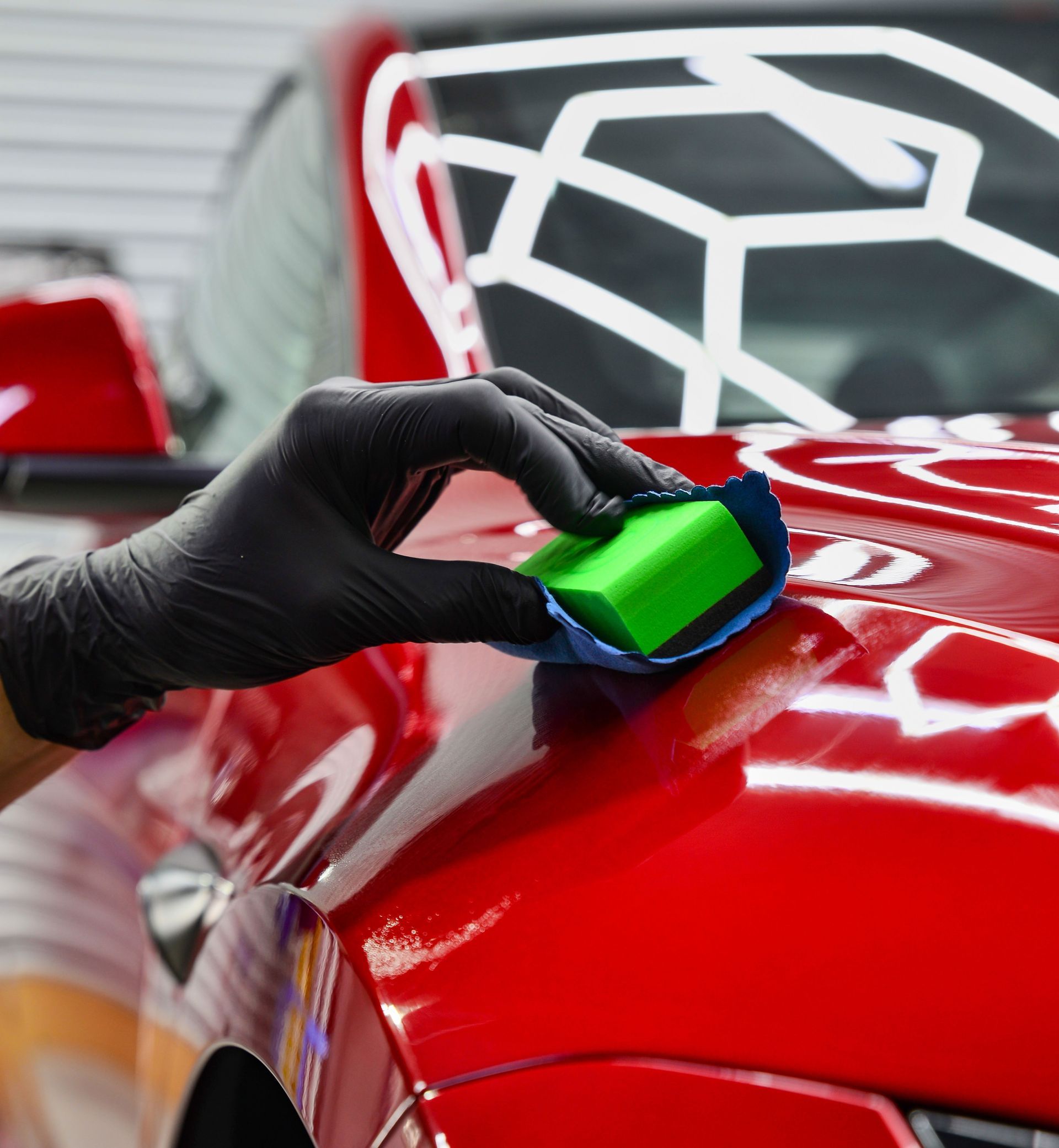 A gloved hand applying car detailing product to a red car with a sponge applicator.