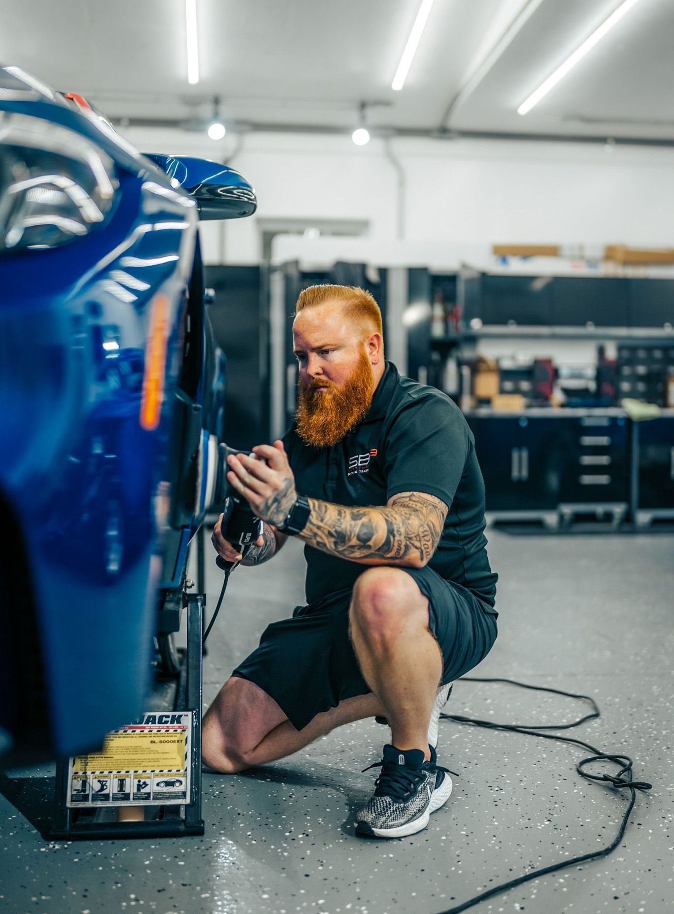 Man with red beard working on blue car wheel in a garage, squatting with tools.