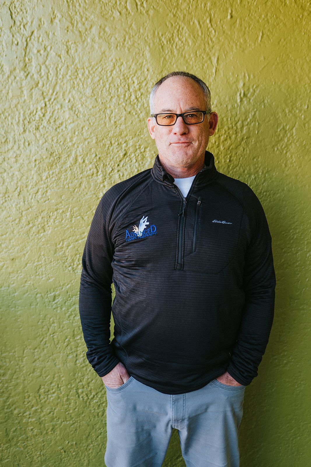 A man wearing glasses and a black sweater is standing in front of a green wall.