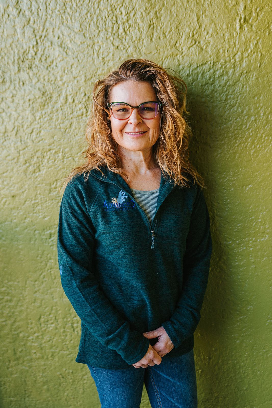 A woman wearing glasses and a green sweater is standing in front of a green wall.