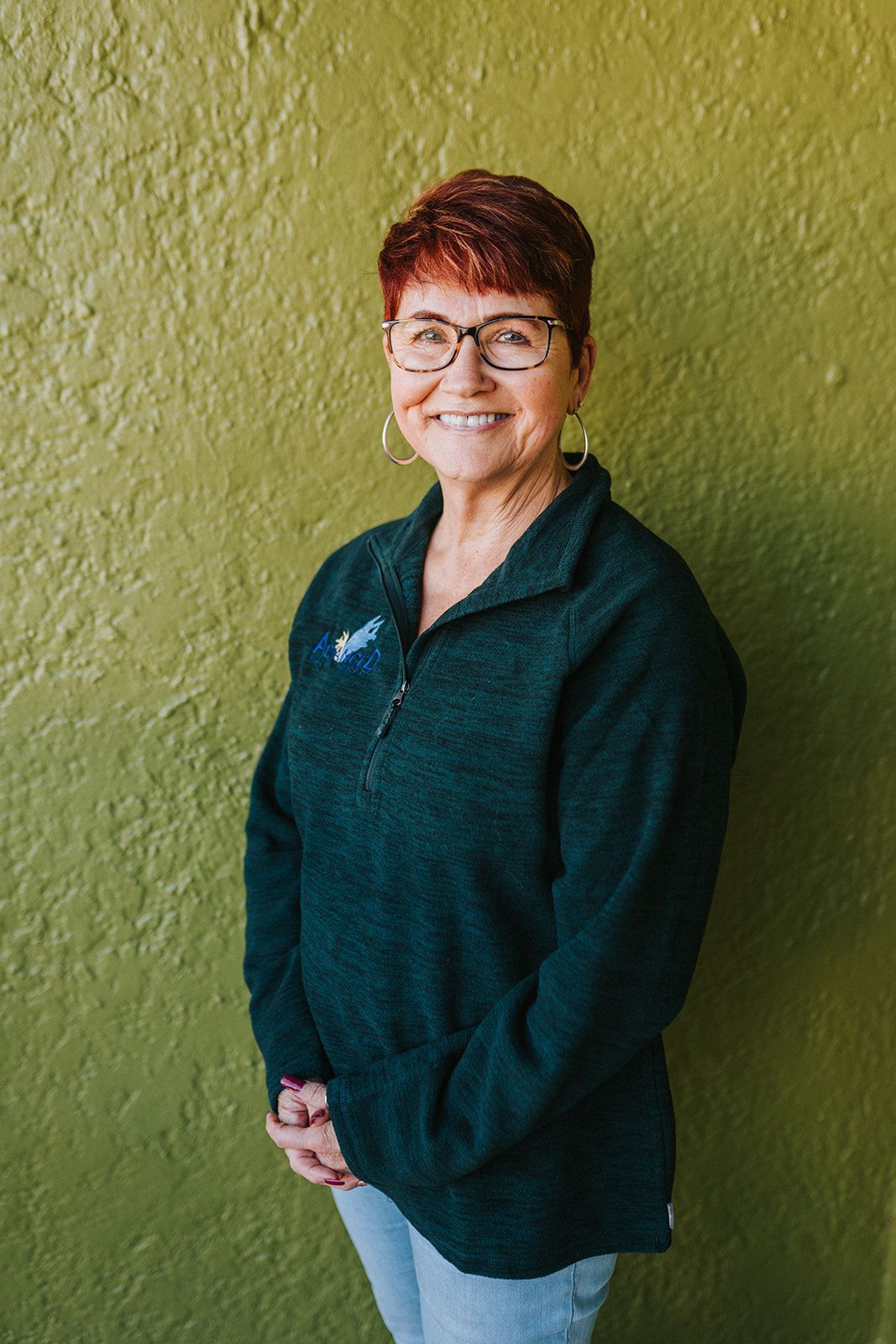 A woman wearing glasses and a green sweater is standing in front of a green wall.