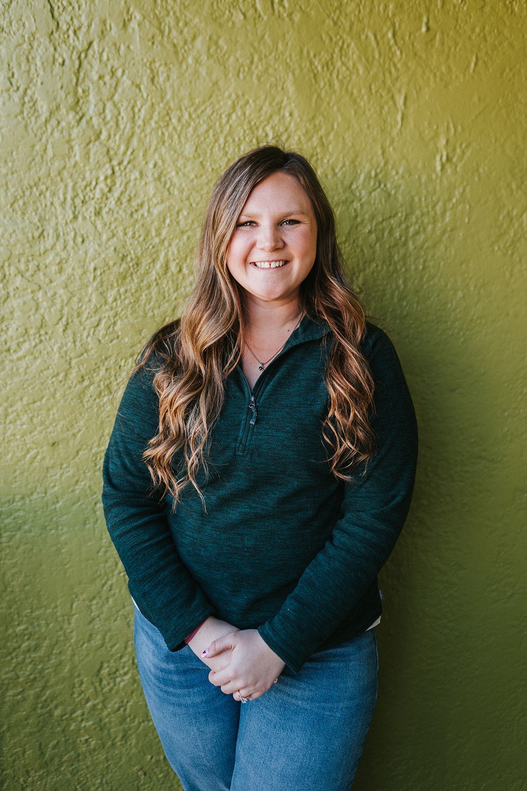 A woman in a green sweater and jeans is standing in front of a green wall.