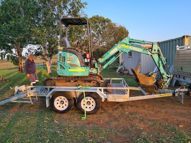 A Man Is Standing Next To A Small Excavator On A Trailer — Rockyview Excavations In Rockyview, QLD