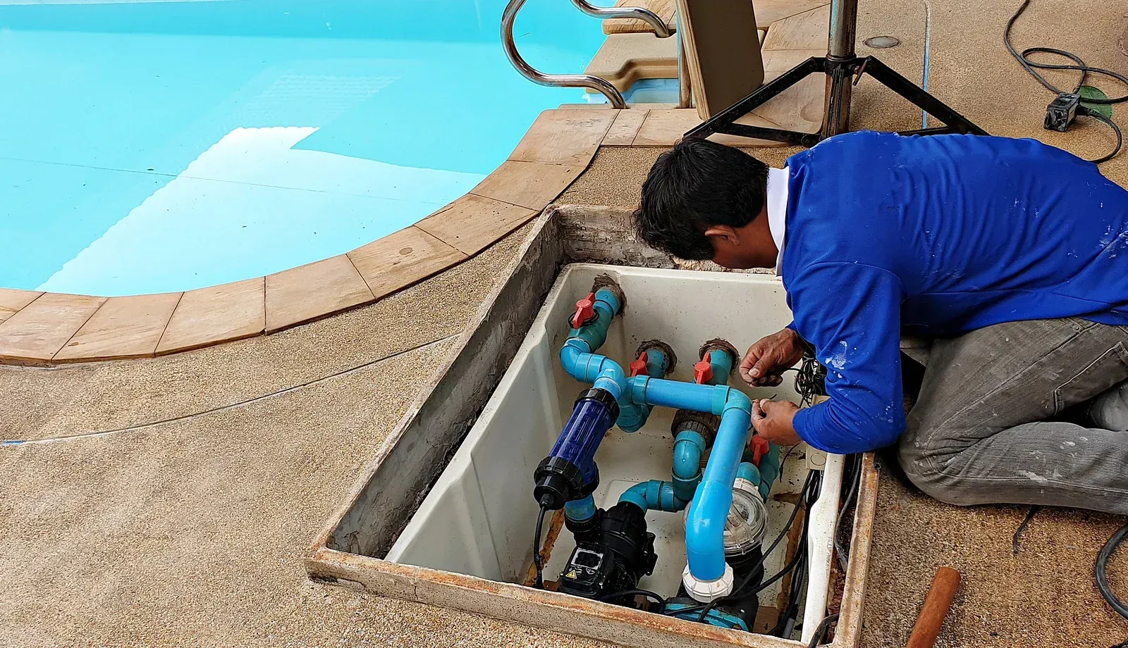 Technician repairing pool plumbing in an open ground access hatch beside a swimming pool