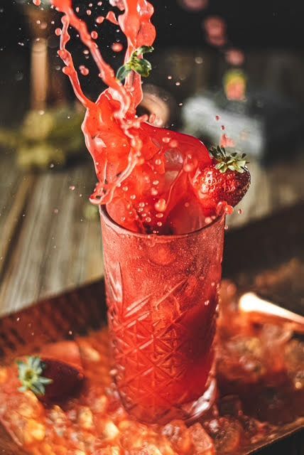 Cóctel de fresas desbordándose de un vaso decorado, con fresas en una bandeja de madera.