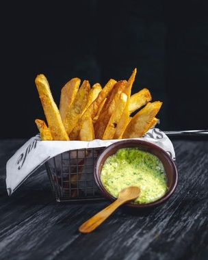 Cesta de patatas fritas sazonadas con salsa verde para mojar, cuchara de madera, sobre una mesa oscura.