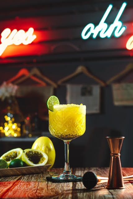 Cóctel amarillo con adorno de lima, maracuyá y un medidor de cobre sobre una barra de madera con luces de neón.