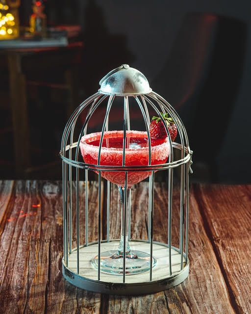 Cóctel servido en una jaula para pájaros, adornado con una fresa, sobre una superficie de madera.