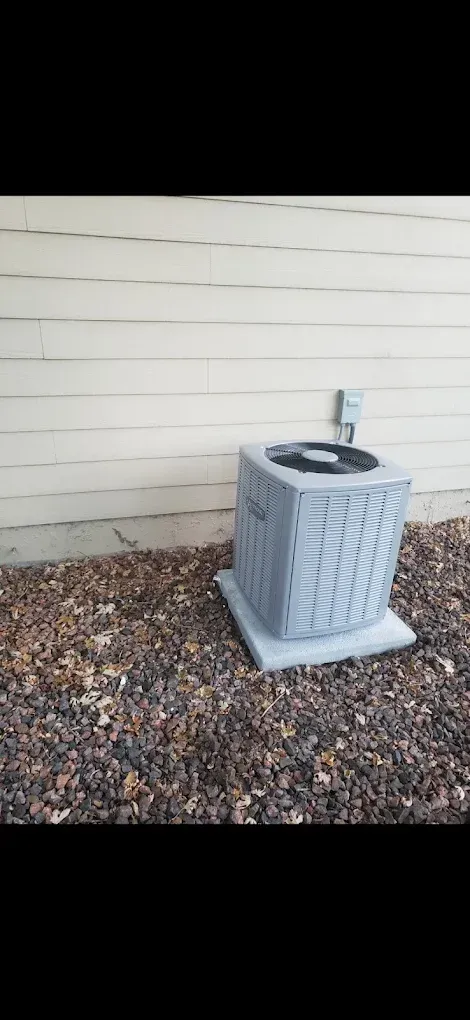 An air conditioner unit sits on a concrete pad next to a beige building. The ground is covered with mulch.