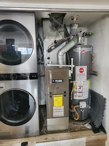 Two technicians in blue overalls inspect a furnace in a home.