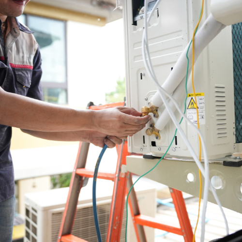 Person working on an air conditioning unit outside. They're on a ladder, connecting a blue hose to the unit.