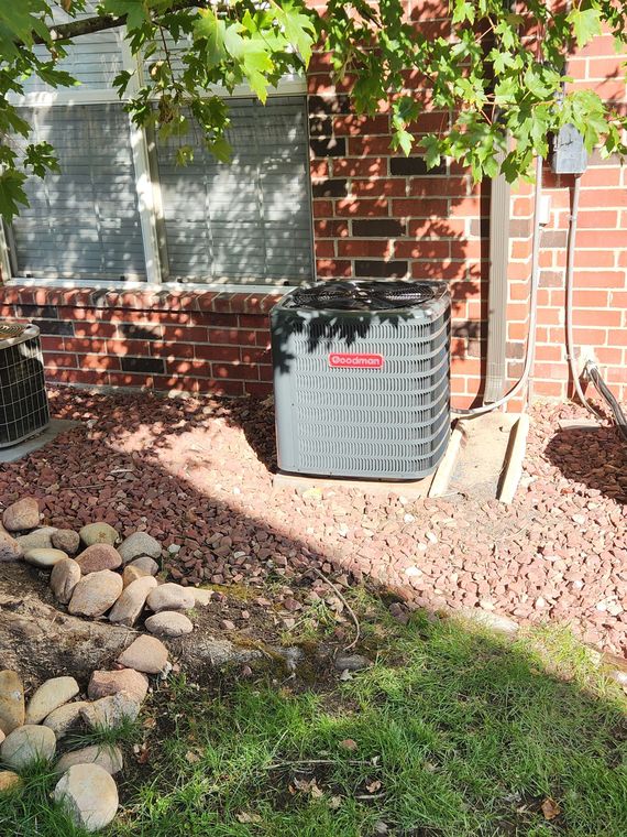 AC unit outside brick building, shaded by a tree, set on red gravel.