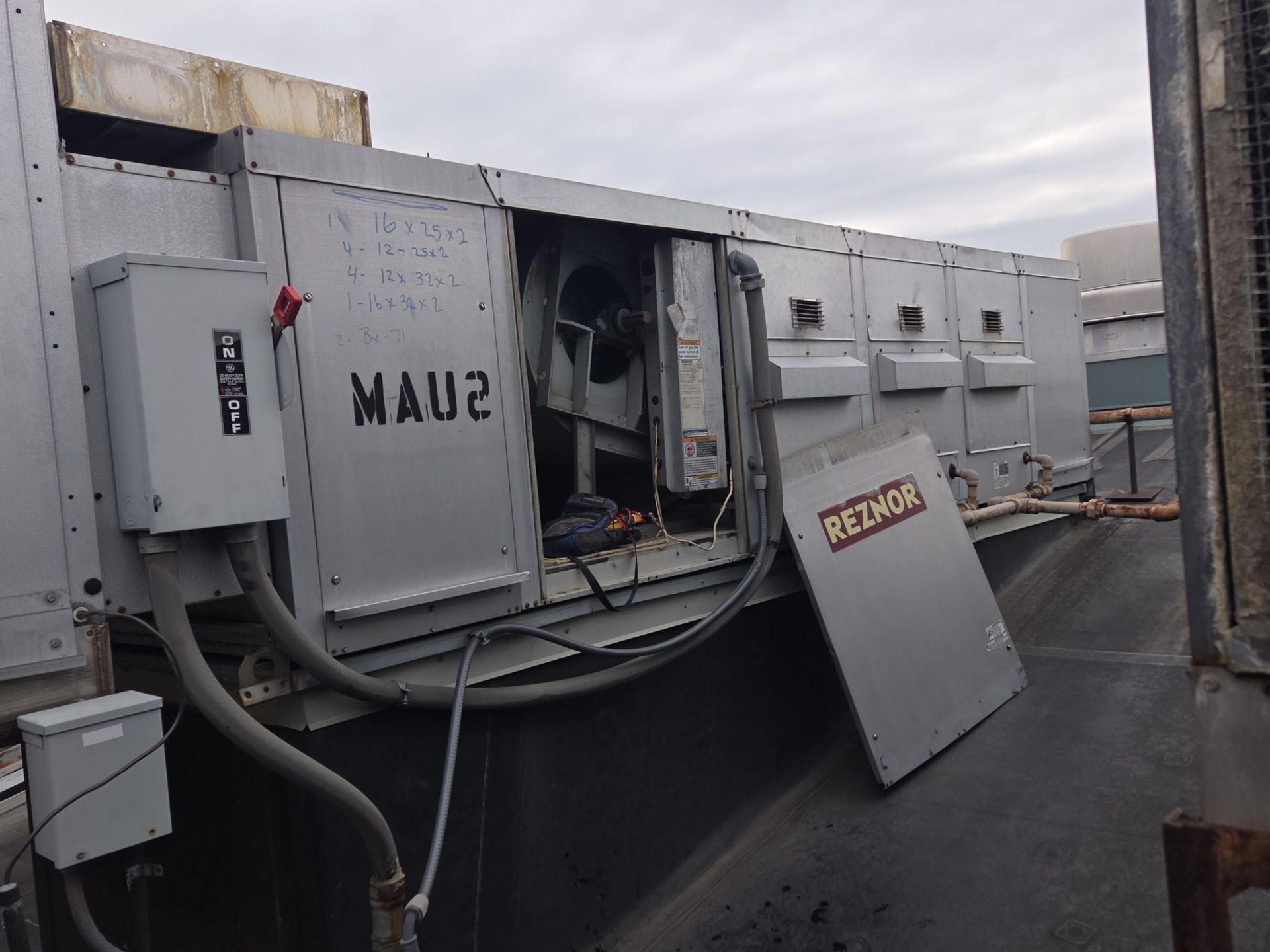 HVAC technician in hardhat, safety vest, working on industrial air conditioning unit. Silver ducts, pipes, building interior.