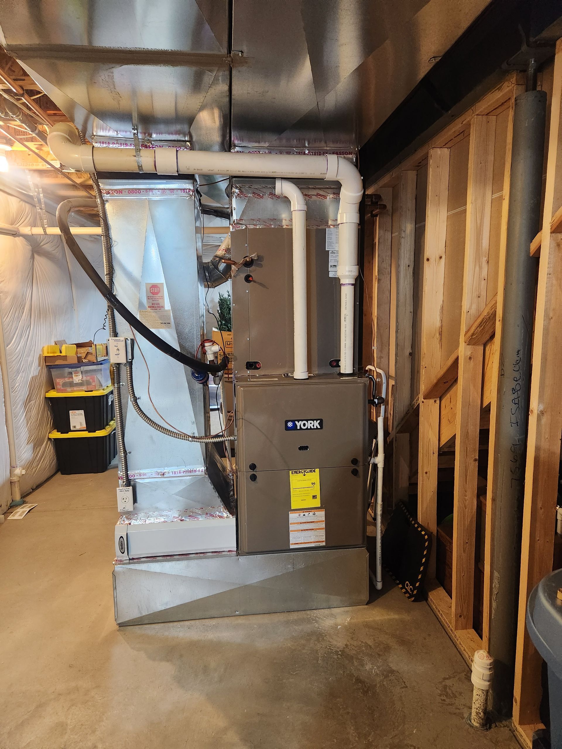 Furnace and ductwork in a basement. Grey furnace, silver ducts, white PVC pipes, and exposed wood studs.