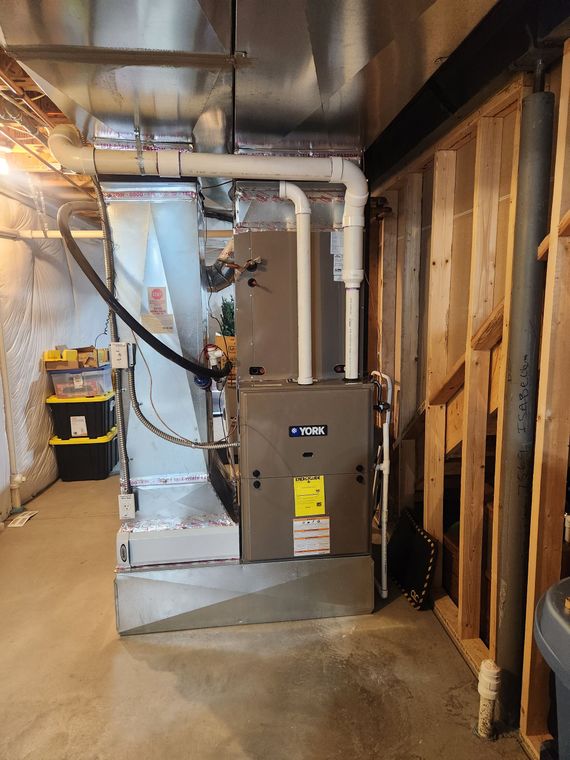 Furnace and ductwork in a basement. Grey furnace, silver ducts, white PVC pipes, and exposed wood studs.