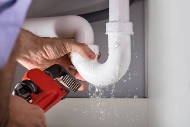 Plumber in blue plaid shirt repairs leaky pipes under a sink; water spills.