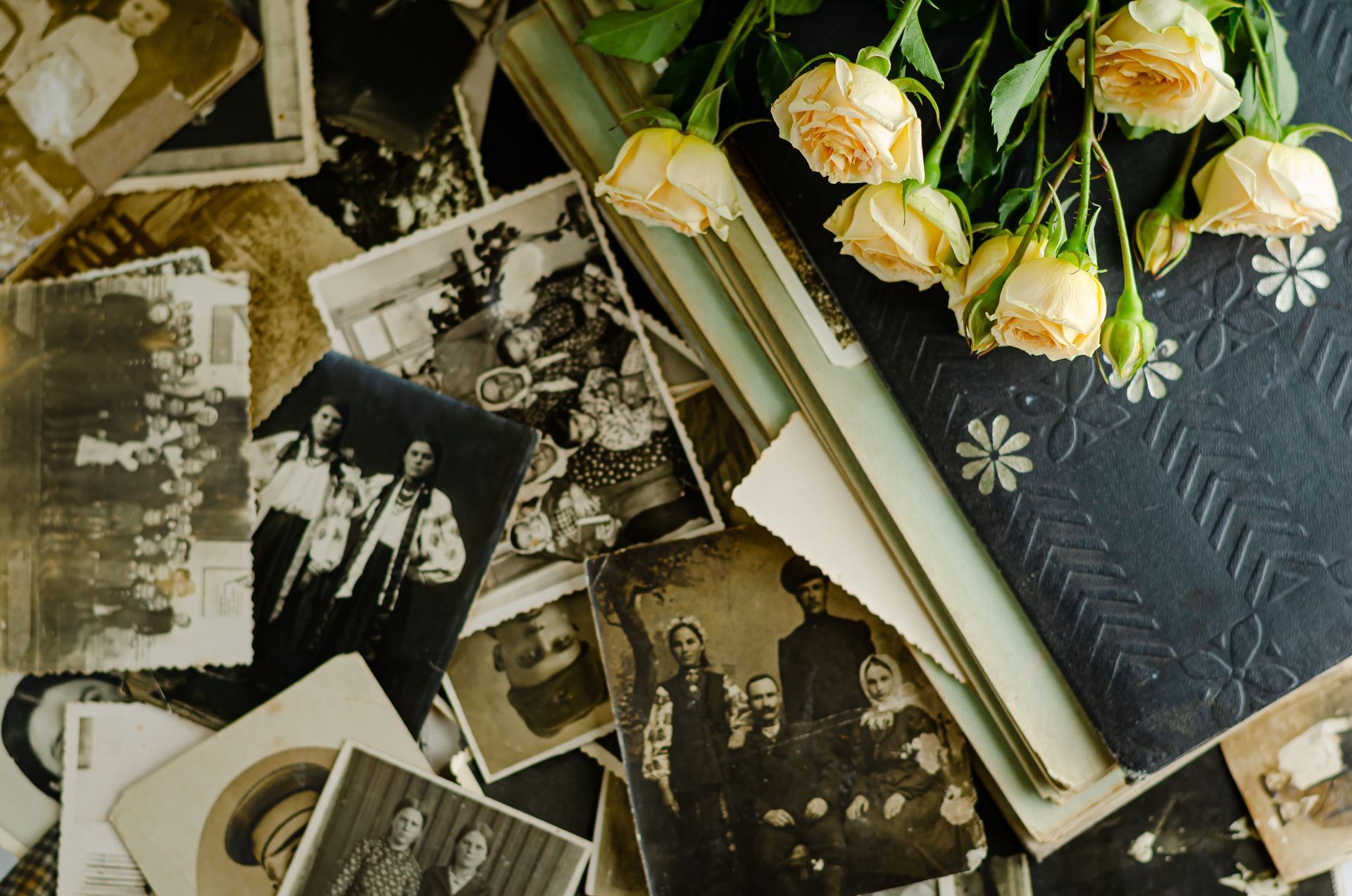 Vintage black-and-white photos scattered on a dark surface next to a bound album and a bouquet of cream-colored roses.