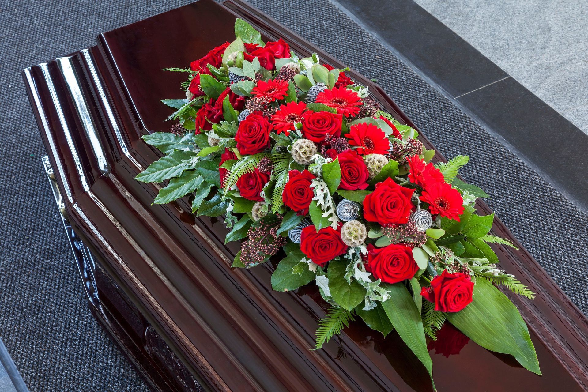 A casket made of dark, polished wood, topped with a lush arrangement of vibrant red roses and green foliage.