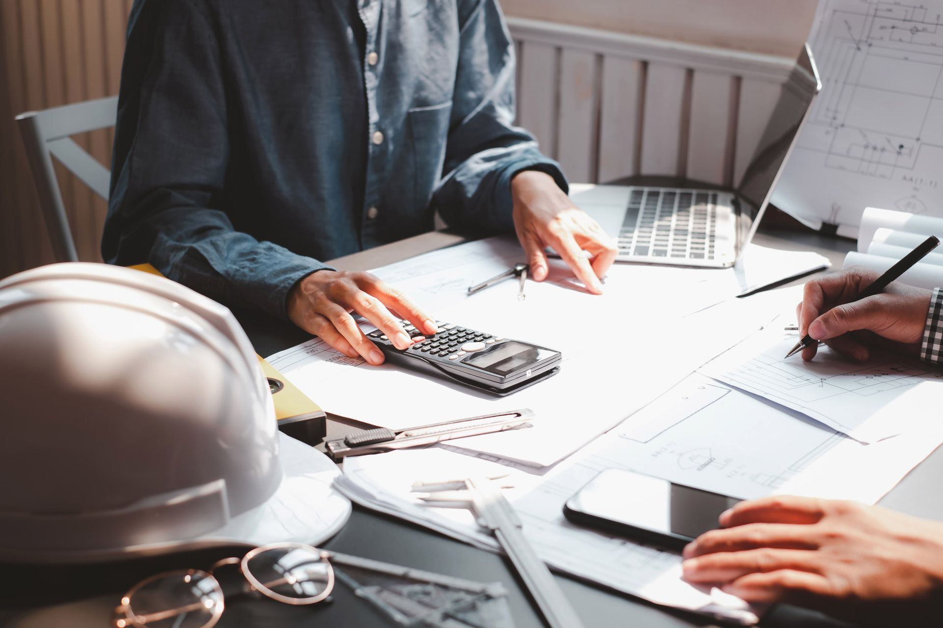 Two people working at a desk with papers, calculator, laptop, and hard hat.
