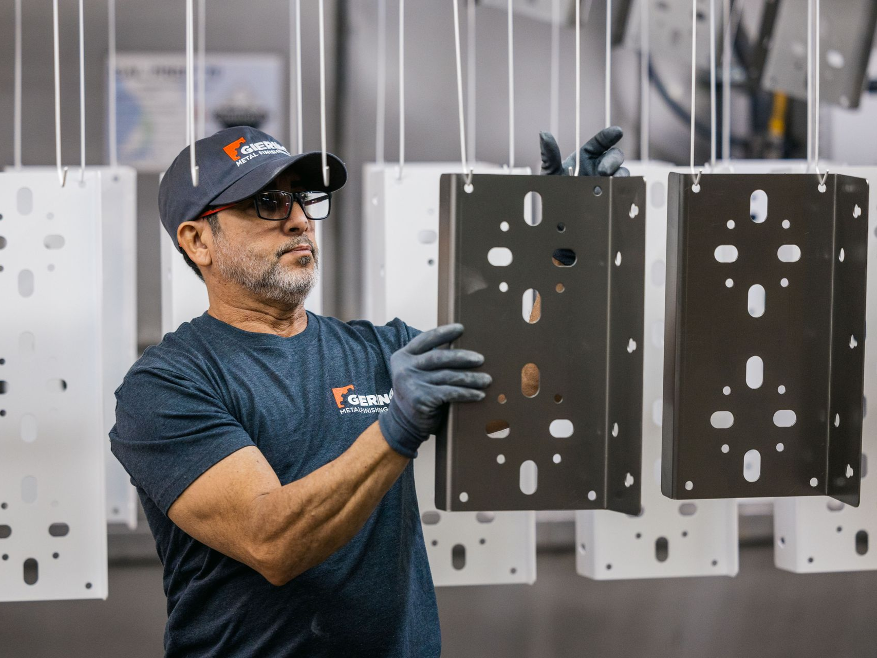 Man inspecting a metal part, wearing gloves and a hat, at a manufacturing facility.