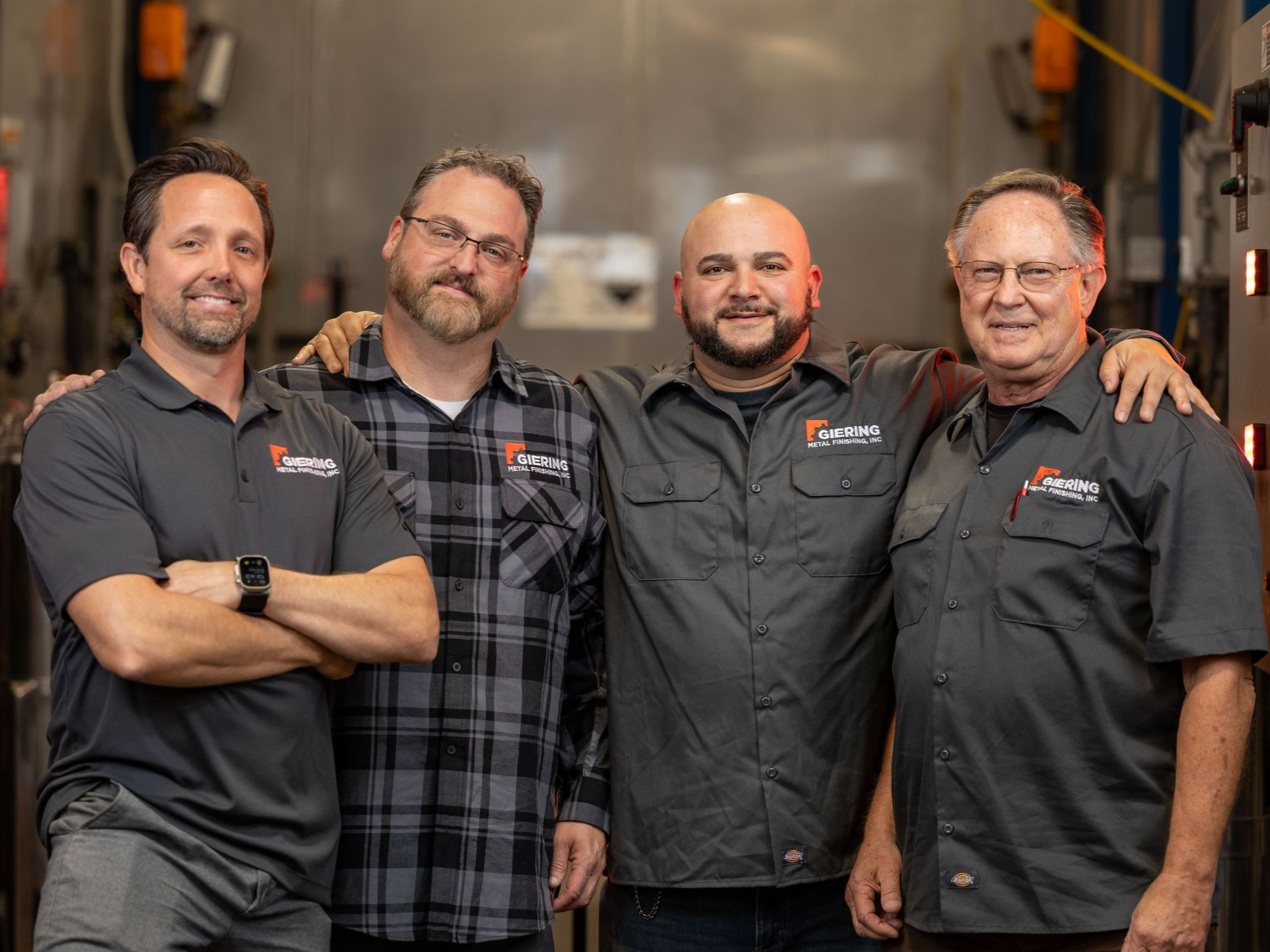 Four men in work shirts stand together, arms around each other, smiling. Industrial setting.